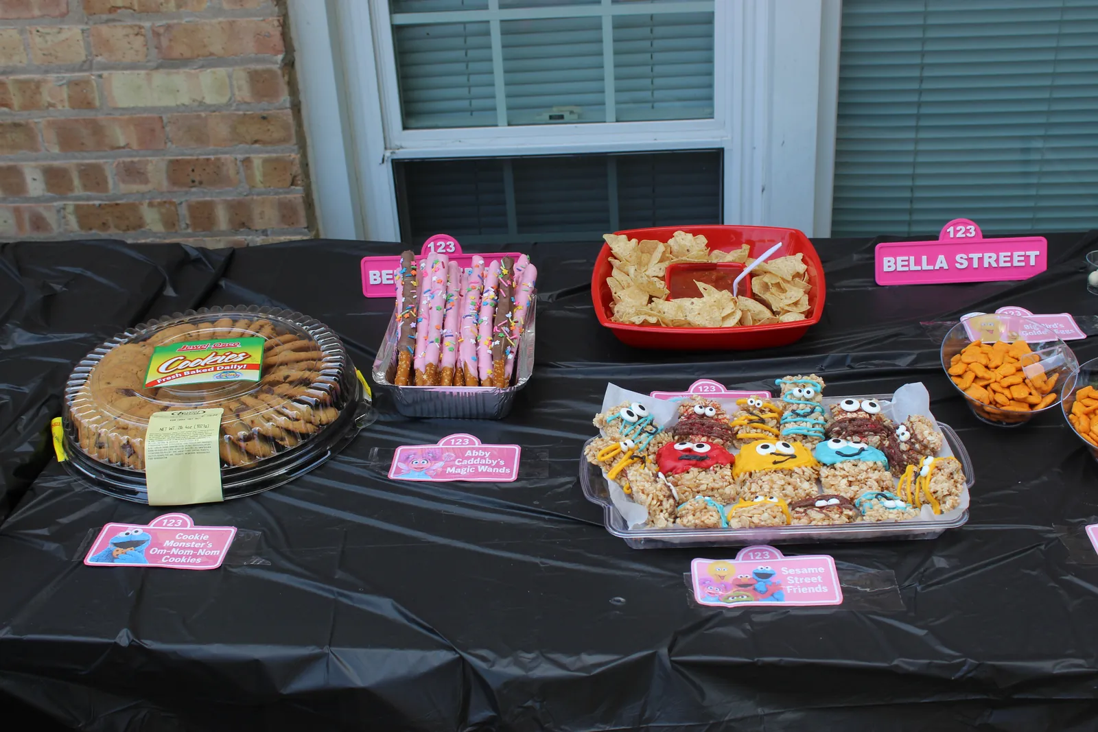 Party table with a cookie tray, chocolate-dipped pretzel rods with pink icing and sprinkles, a red bowl of tortilla chips and salsa, and rice krispie treats decorated as Sesame Street characters, each behind a small pink card reading Abby Cadabby’s Magic Wands and Sesame Street Friends