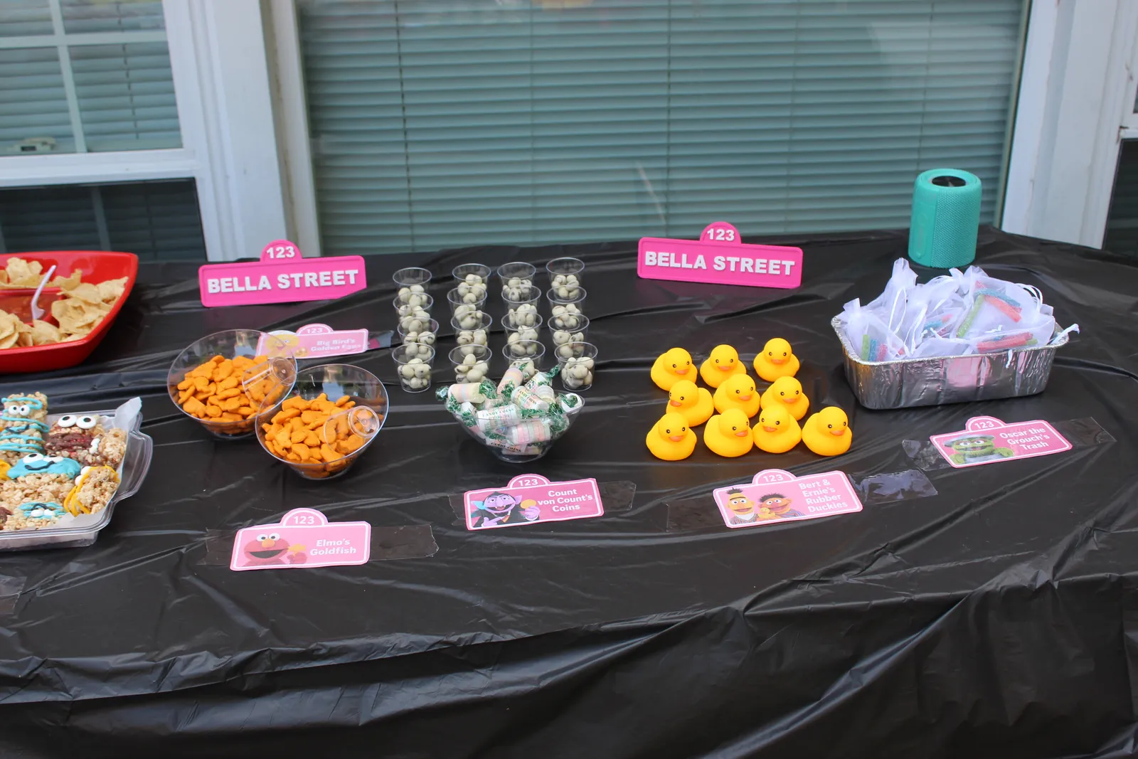 Close view of a black-covered party table with goldfish crackers in clear bowls, cups of candy, wrapped candy coins, nine yellow rubber ducks, and a foil tray of favor bags, each behind a small pink Sesame Street card reading Elmo’s Goldfish, Count von Count’s Coins, Bert and Ernie’s Rubber Duckies, and Oscar the Grouch’s Trash