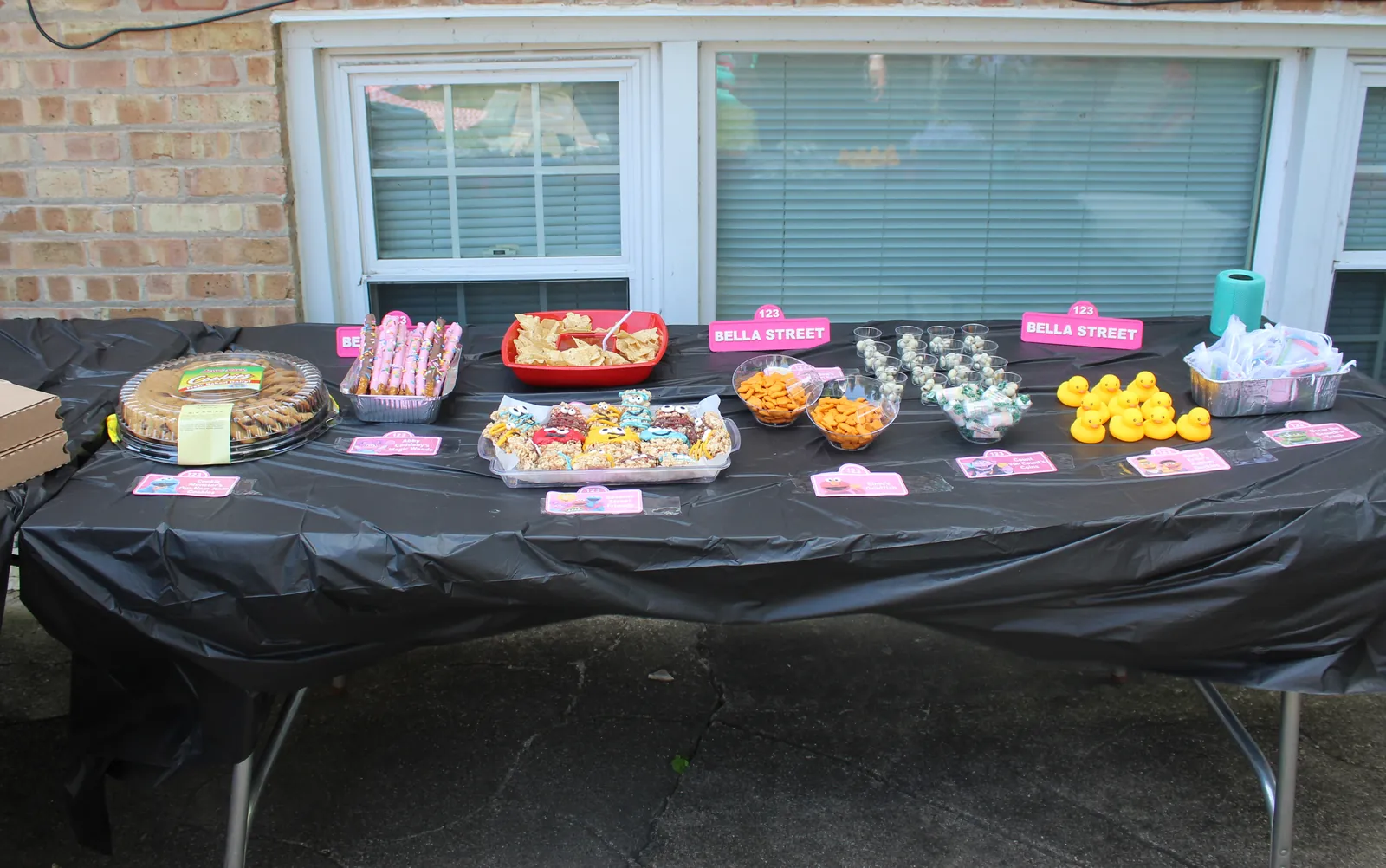 Long party table under a brick wall, covered in black plastic and set with cookies, chocolate-dipped pretzels, tortilla chips, decorated rice krispie treats, goldfish crackers, candy cups, and rubber ducks, with pink 123 BELLA STREET signs standing between them and small pink labeled cards in front of each dish